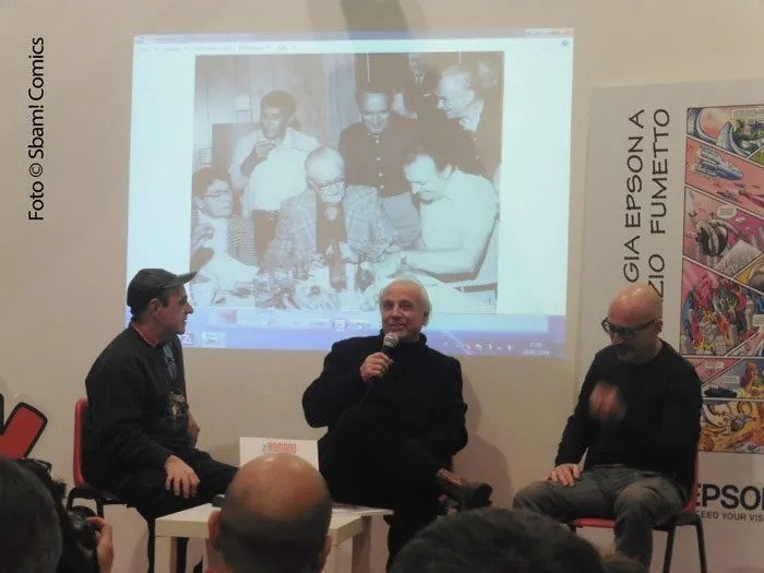 Da sinistra: Luca Boschi, Marco Rota e Davide Catenacci; sopra di loro, la foto dello storico incontro tra Carl Barks (seduto al centro, con la giacca a quadri) che si scambia disegni con Romano Scarpa (a destra)